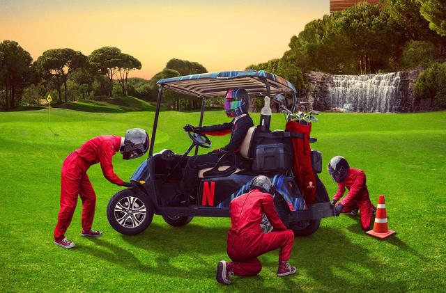 A person wearing a Formula 1 helmet sits in a golf buggy on a golf course as technicians swap the wheels.