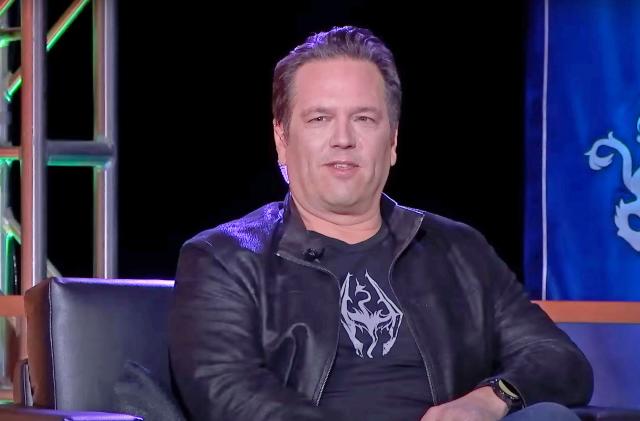 Xbox's Phil Spencer is shown sitting down on stage at an event. The photo is closely cropped so you only see him and the edges of his black chair.