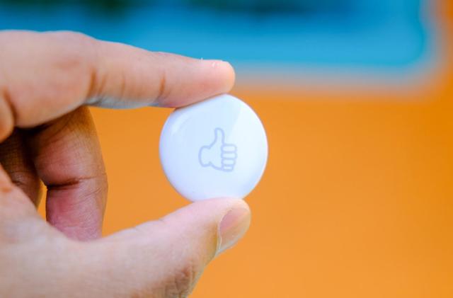 A white Apple AirTag Bluetooth tracker with a "thumbs up" emoji engraved on the front of it is held in between a person's thumb and forefinger.