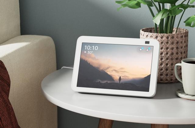 A white Amazon Echo Show 8 displays the time and temperature while resting on a side table in front of a potted plant and cup of coffee, next to a white couch.