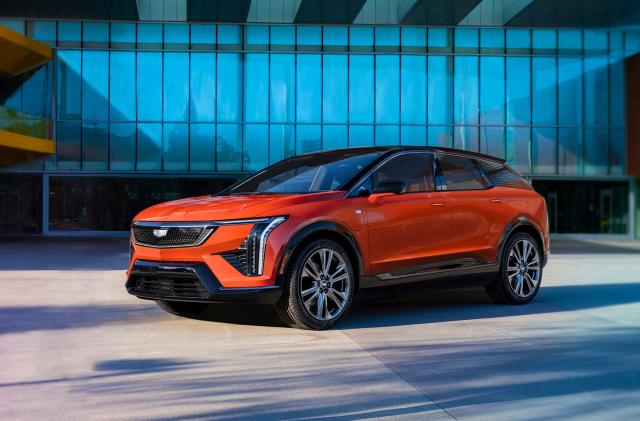 Marketing photo for the Cadillac Optiq. It shows the compact SUV in orange sitting in front of a modern luxury home or garage.