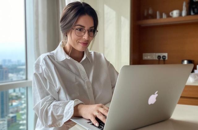 A woman sits at a table on her silver MacBook. 