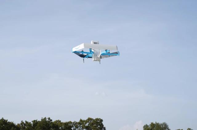 Amazon marketing photo of a Prime Air drone (mostly white with blue accents) flying in the sky. Tops of trees visible below.