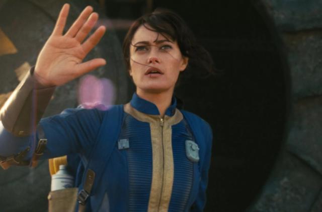 Still from the upcoming "Fallout" TV series. Ella Purnell ("Yellowjackets") stands waving with a hesitant look on her face. She wears a blue uniform with gold trim.