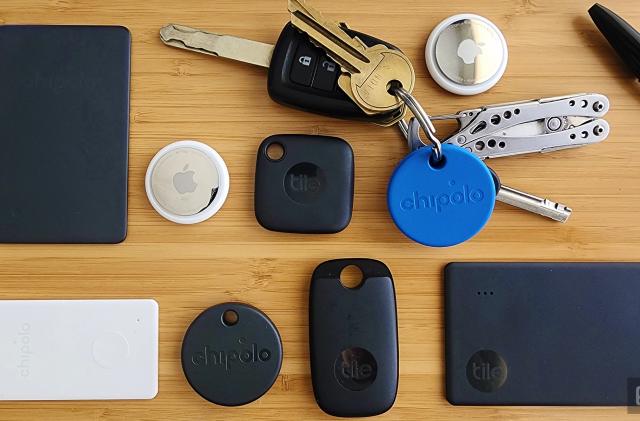 An assortment of bluetooth trackers arranged in a grid on a wooden background. Trackers include black Tile trackers in various shapes, two silver and white AirTag trackers and a round blue Chipolo tracker attached to a set of keys with a multitool key chain. 