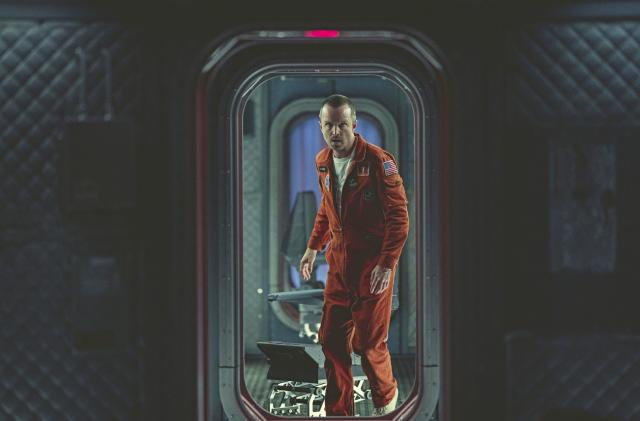 In this press image for Black Mirror, Aaron Paul wears an orange jumpsuit inside the interior of a spaceship (apparently) and looks through a portal doorway.