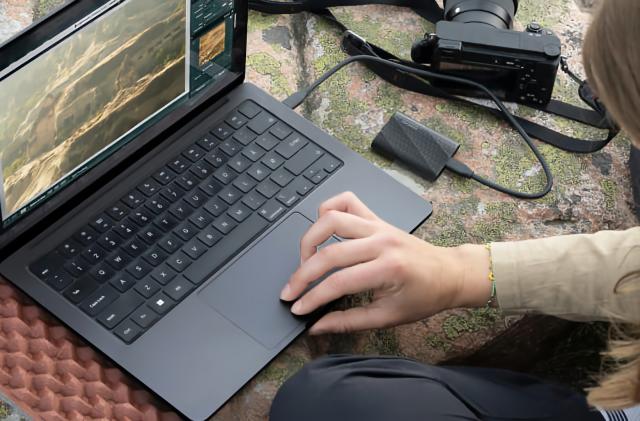 Lifestyle marketing photo of a person using a Windows laptop with a Samsung portable SSD plugged into it. A camera is also nearby, and it all sits on a green and orange surface.