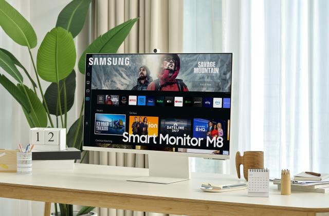 A press shot displaying the Samsung Smart Monitor M8 on a light brown wooden table next to a potted houseplant and an assortment of desktop accessories.