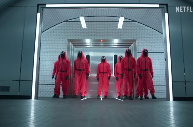 People dressed as Squid Game guards (reddish-pink suits with black faces) stand in a cold industrial building.